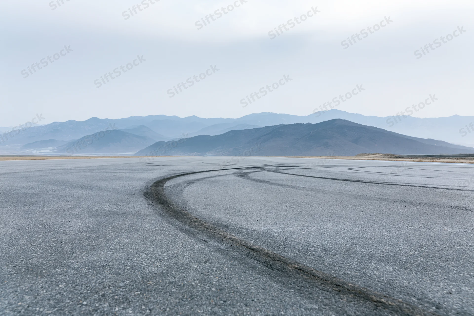 Curved asphalt racetrack with dark rubber burnout marks leading toward distant mountains under overcast sky