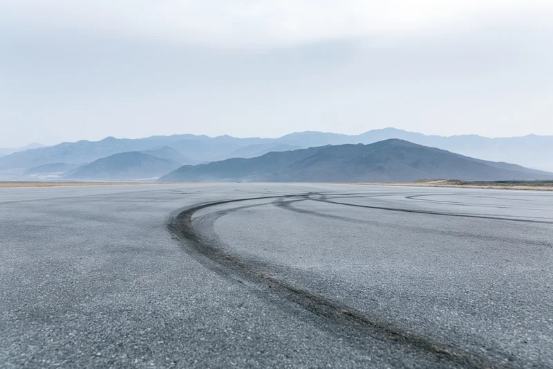 Curved asphalt racetrack with dark rubber burnout marks leading toward distant mountains under overcast sky