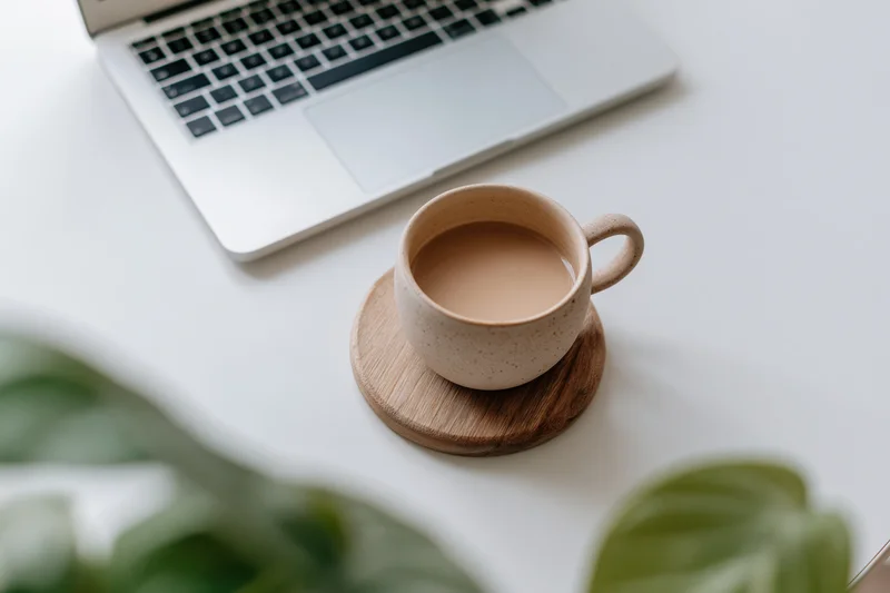 Ceramic mug with coffee on wooden coaster beside silver laptop on white desk with green leaves in foreground