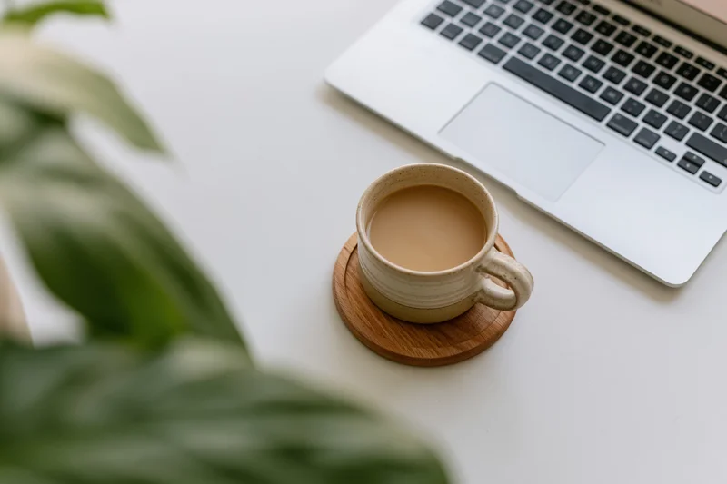 Ceramic mug of coffee on wooden coaster beside silver laptop on white desk with green plant leaves in foreground