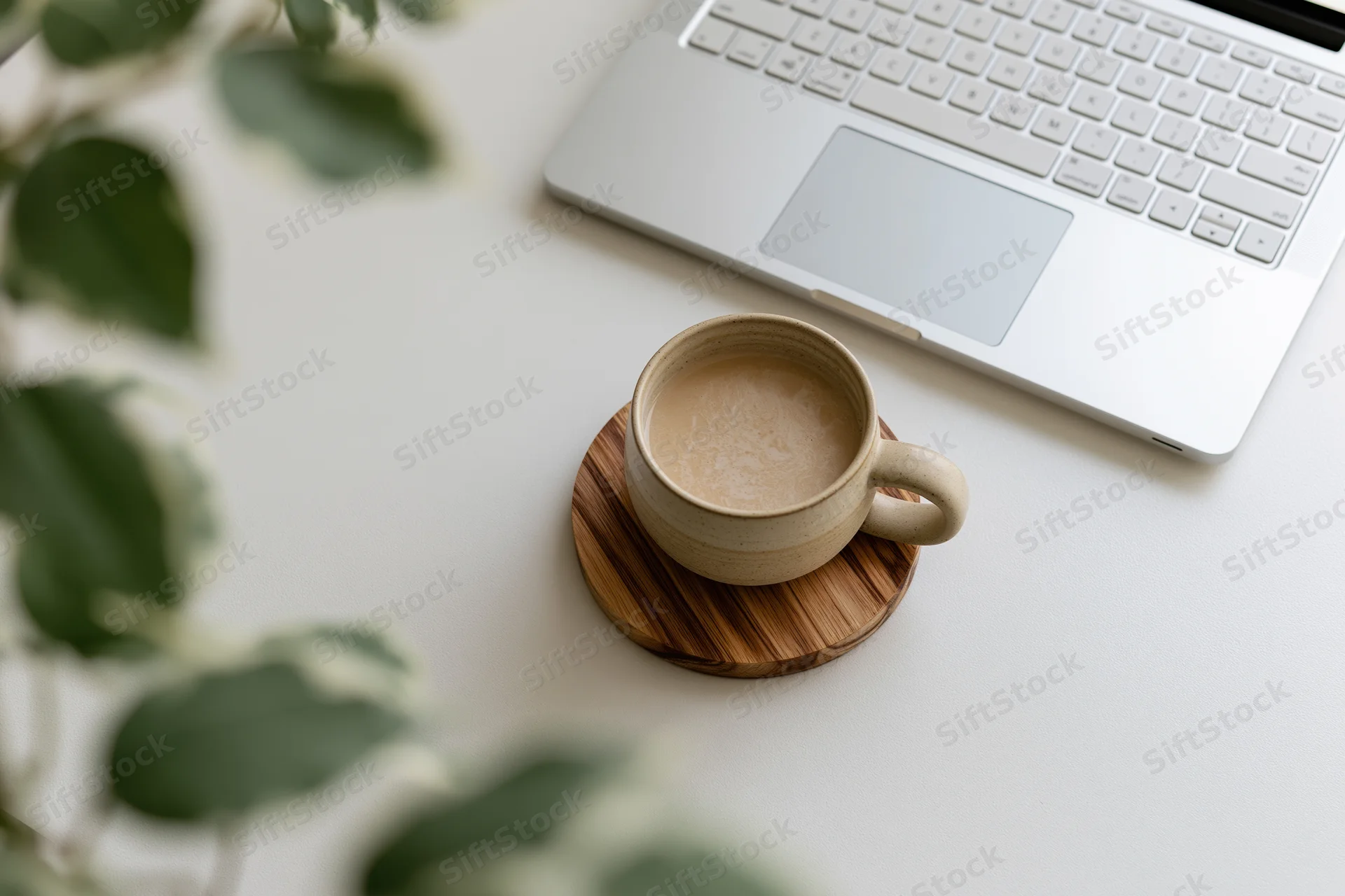 Ceramic coffee mug on bamboo coaster beside silver laptop on white desk with green plant leaves in foreground