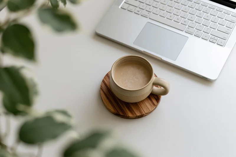 Ceramic coffee mug on bamboo coaster beside silver laptop on white desk with green plant leaves in foreground