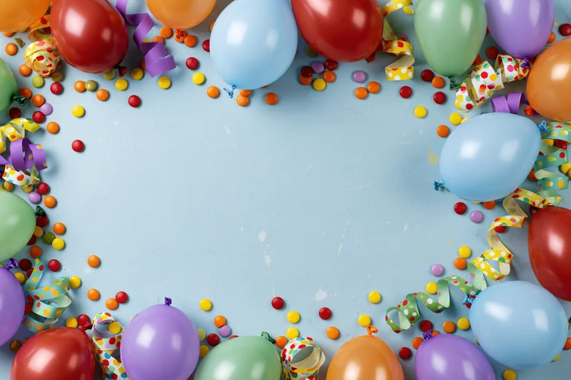 Colorful party balloons and streamers arranged in a border around a light blue surface with confetti and copyspace in the center