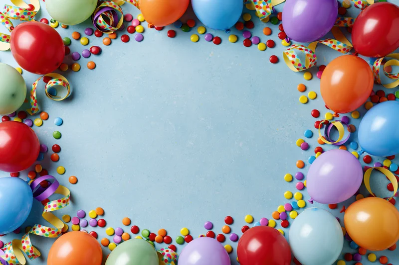 Colorful party balloons and streamers arranged in a frame around a blue surface with confetti and copyspace