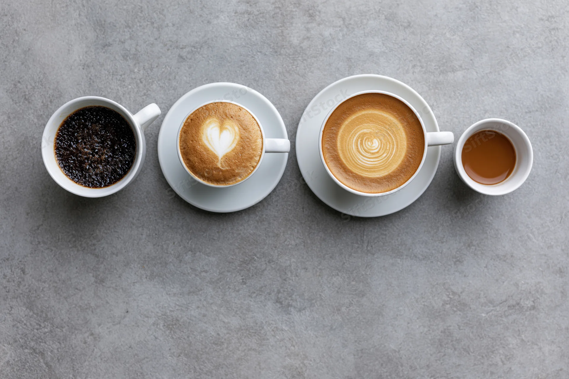 Four white ceramic coffee cups in a row with different coffee types and latte art on a gray surface, overhead view