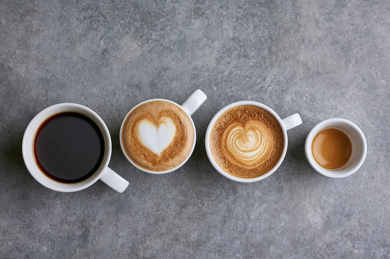 Four white ceramic coffee cups in a row on a gray surface, each filled with a different coffee beverage from black coffee to espresso with latte art