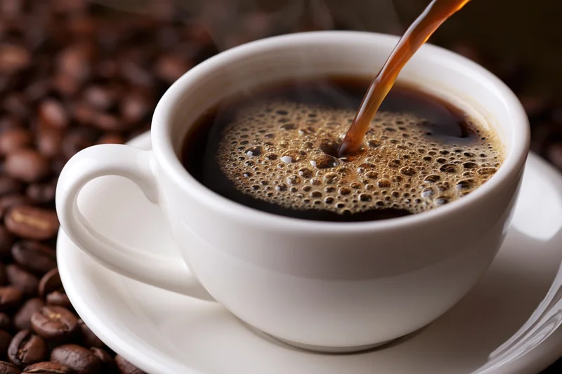 Hot black coffee being poured into a white ceramic cup on a saucer surrounded by roasted coffee beans