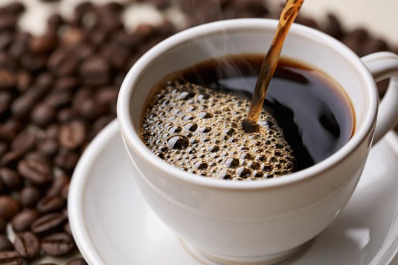 Hot black coffee being poured into a white ceramic cup with steam and bubbles, surrounded by roasted coffee beans on a saucer