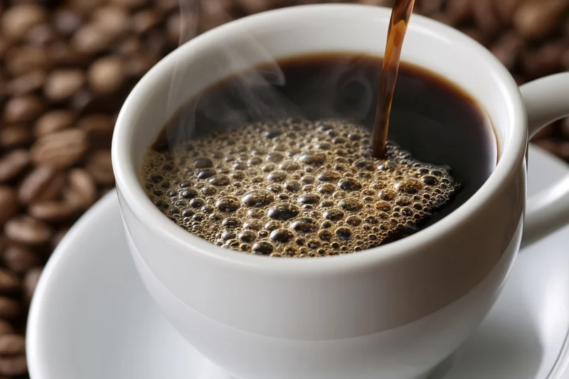 Hot black coffee being poured into a white ceramic cup with steam and bubbles, set on a saucer over coffee beans