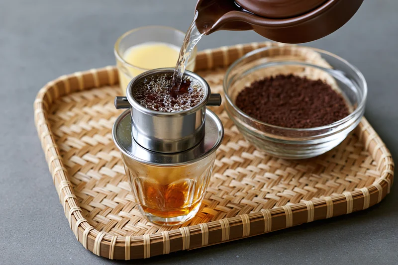 Hot water being poured into a Vietnamese phin coffee filter on a woven bamboo tray