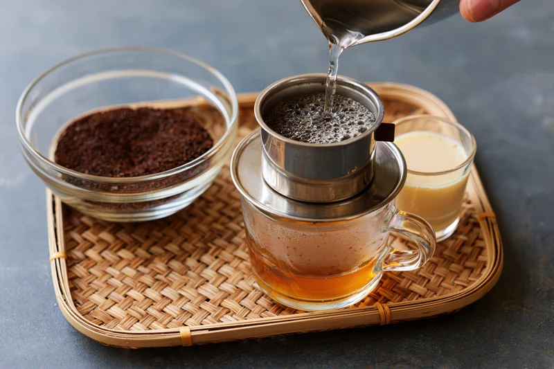 Hand pouring hot water into a Vietnamese phin filter over a glass mug on a woven tray