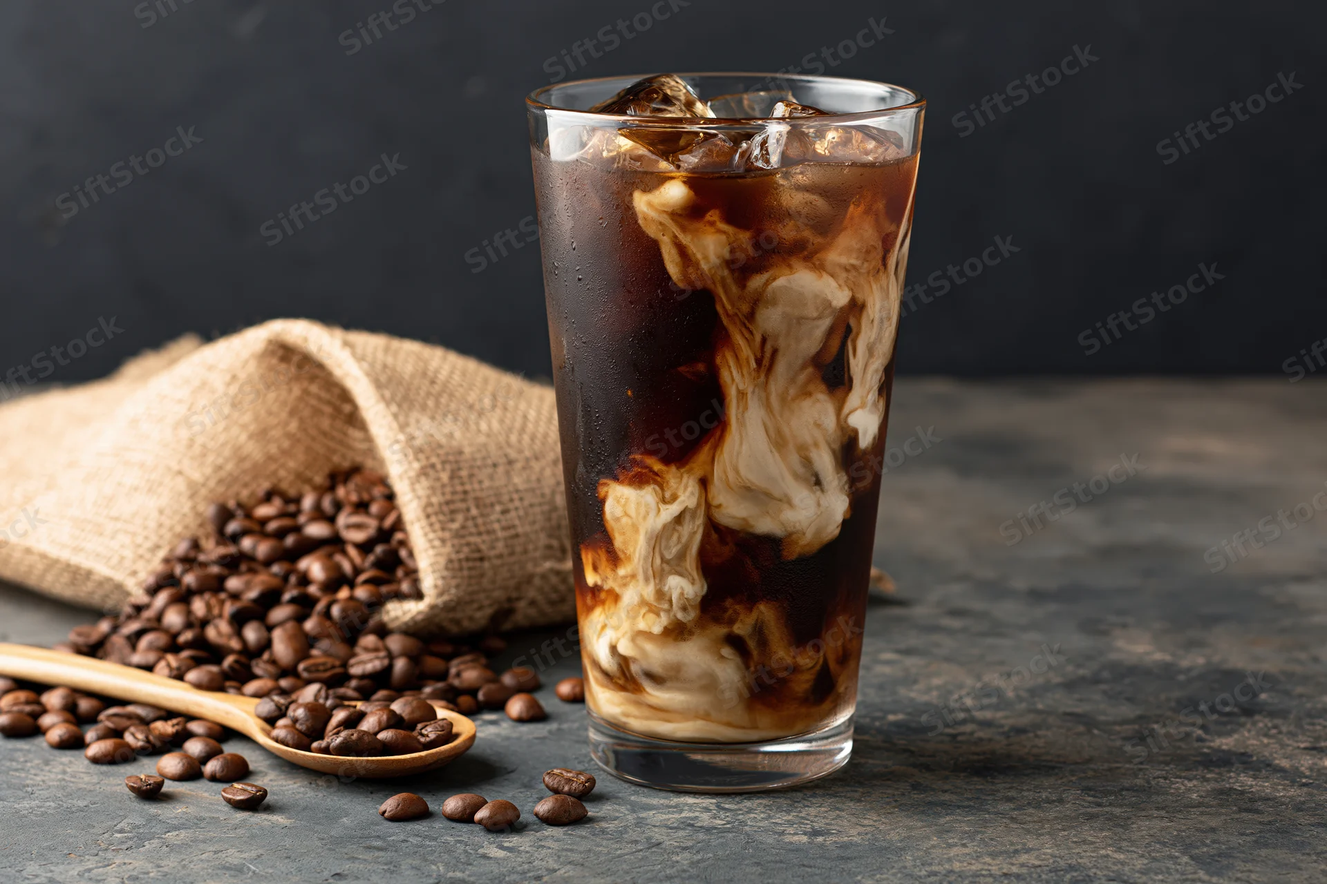 Iced coffee with swirling cream in a tall glass beside coffee beans and a burlap sack on a stone surface