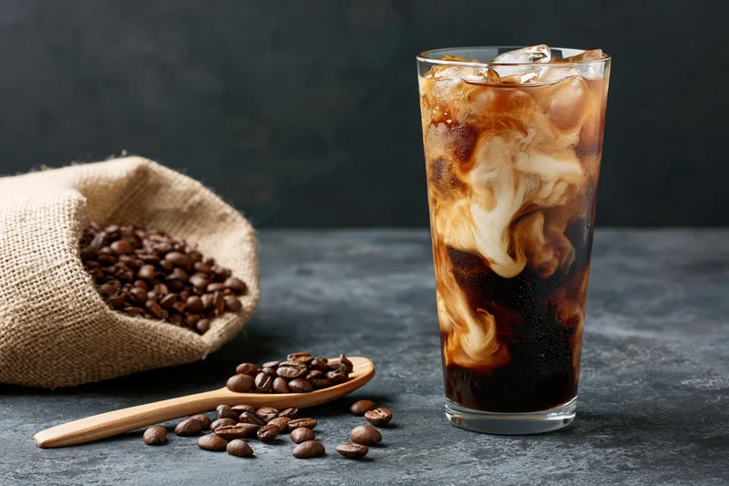 Iced coffee with swirling cream in a tall clear glass beside coffee beans in a burlap sack and on a wooden spoon