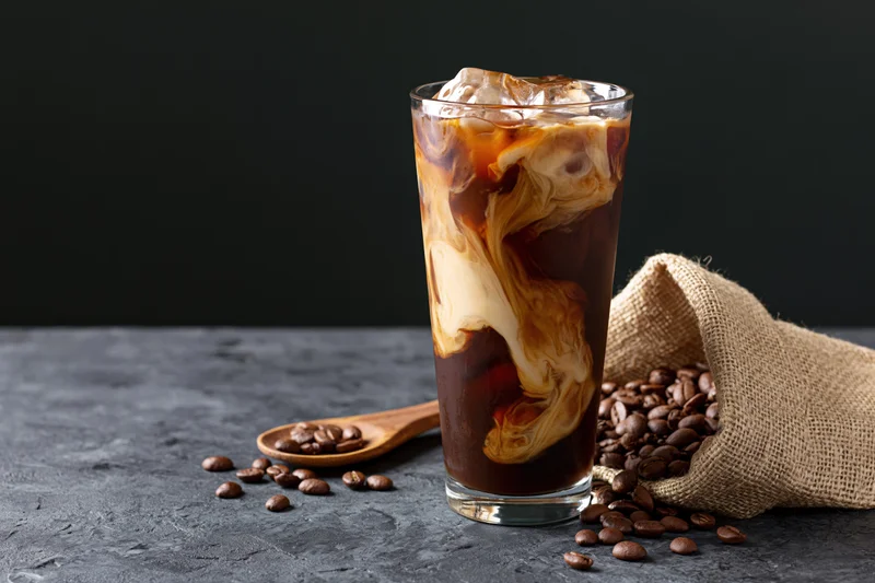 Iced coffee with swirling cream in a tall glass on a dark stone surface beside coffee beans and a burlap sack