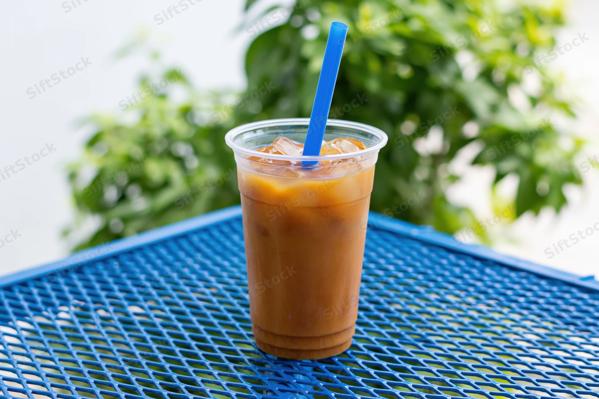 Iced coffee in a clear plastic cup with blue straw on a blue grid table outdoors