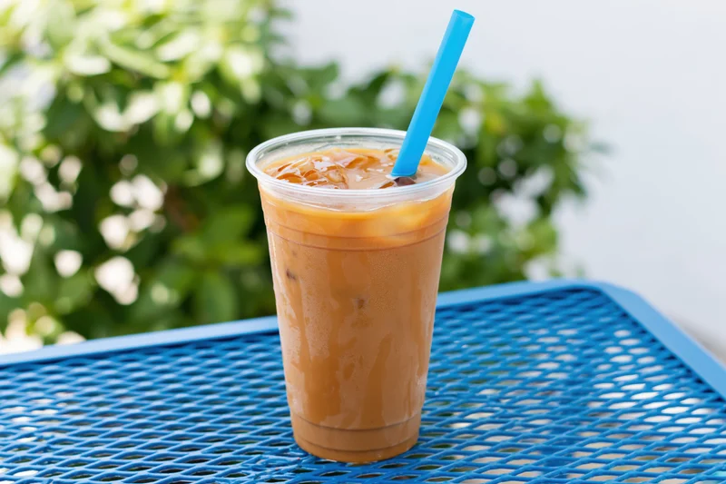 Iced milk tea in a clear plastic cup with blue straw on a blue mesh table outdoors