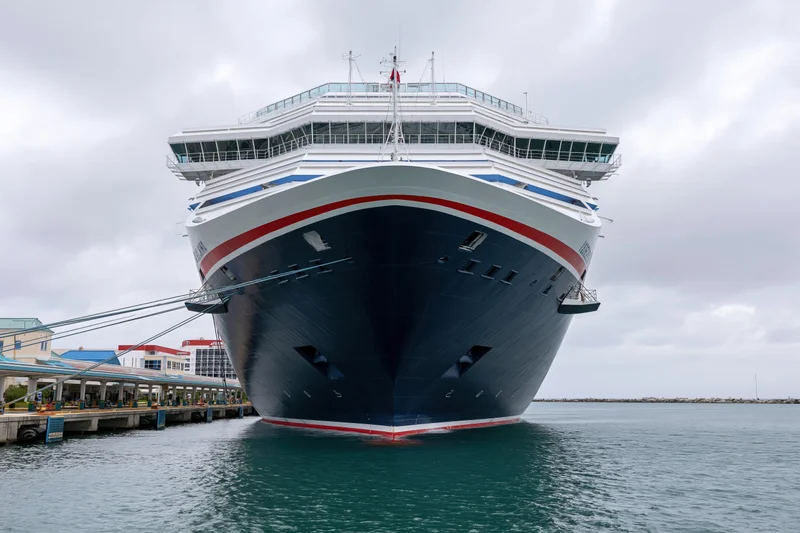 Large modern cruise ship docked at port with bow facing camera, moored to pier under cloudy sky