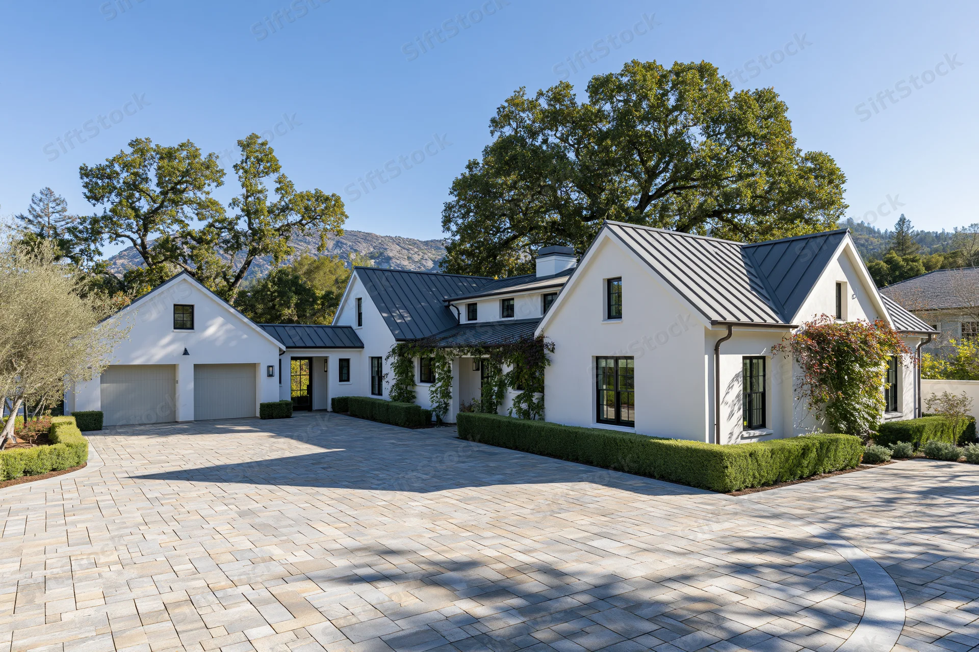Modern white stucco farmhouse with dark metal roof and stone driveway surrounded by trees and mountains