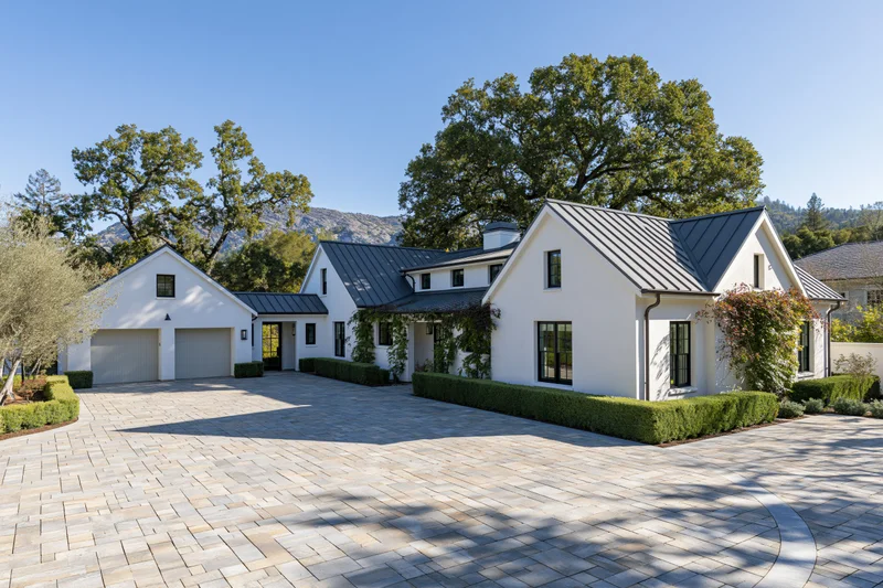 Modern white stucco farmhouse with dark metal roof and stone driveway surrounded by trees and mountains