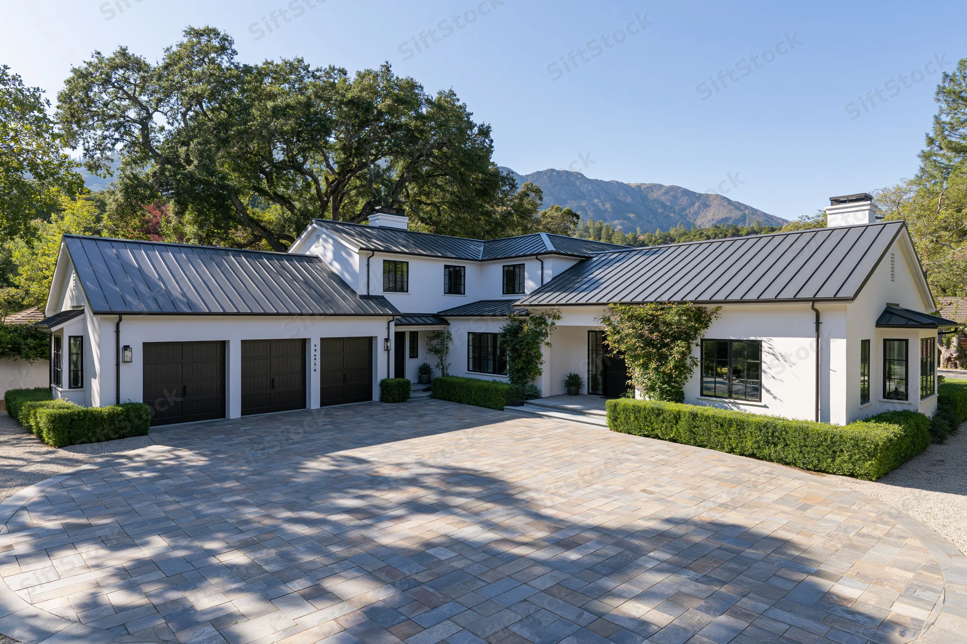 Modern white stucco farmhouse with dark metal roof and stone driveway surrounded by trees and mountains