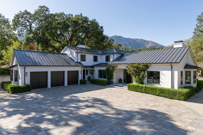 Modern white stucco farmhouse with dark metal roof and stone driveway surrounded by trees and mountains