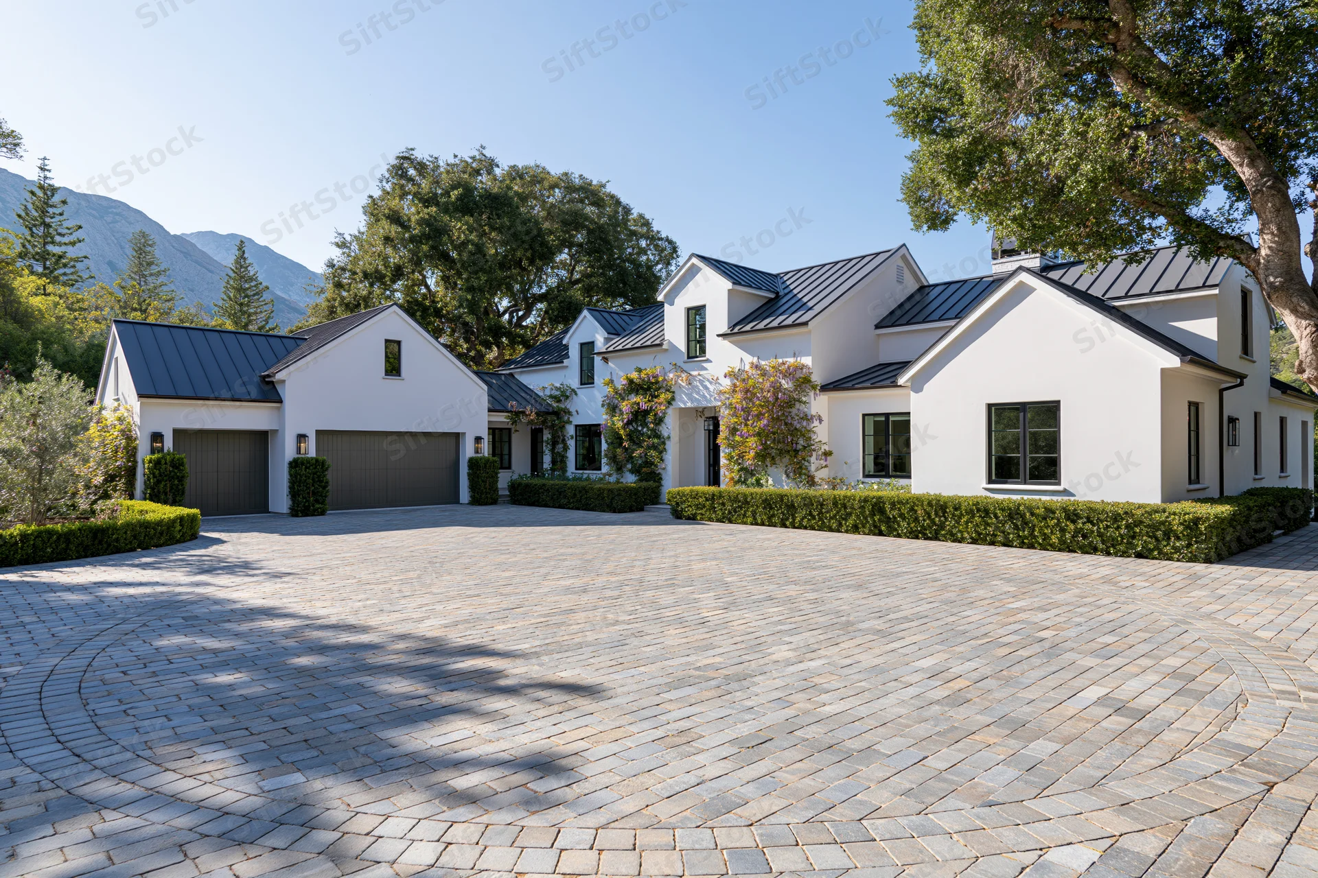 Modern white stucco farmhouse with dark metal roof and circular cobblestone driveway surrounded by trees and mountains