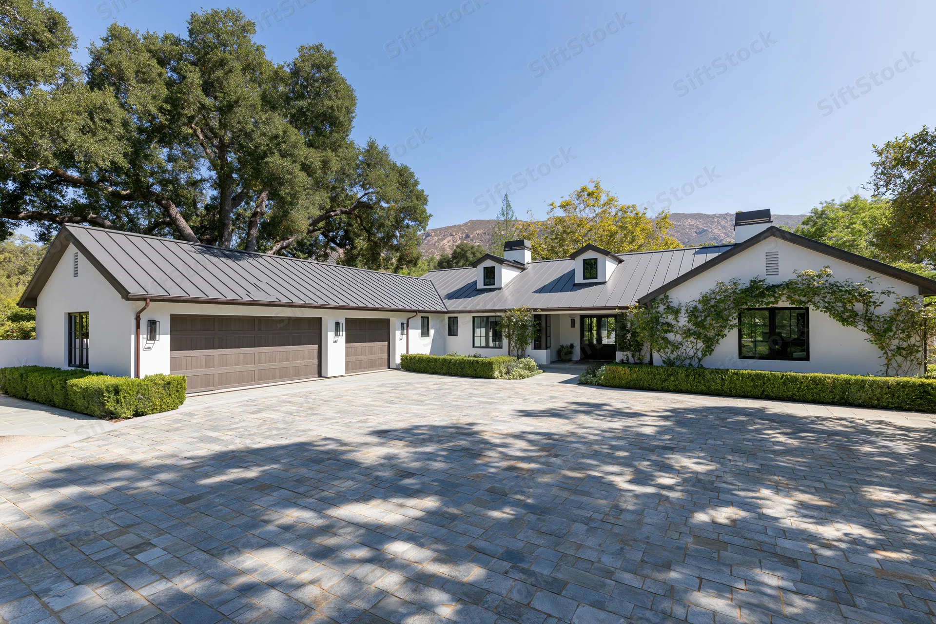 Modern white stucco farmhouse with dark metal roof and stone driveway surrounded by trees and hills