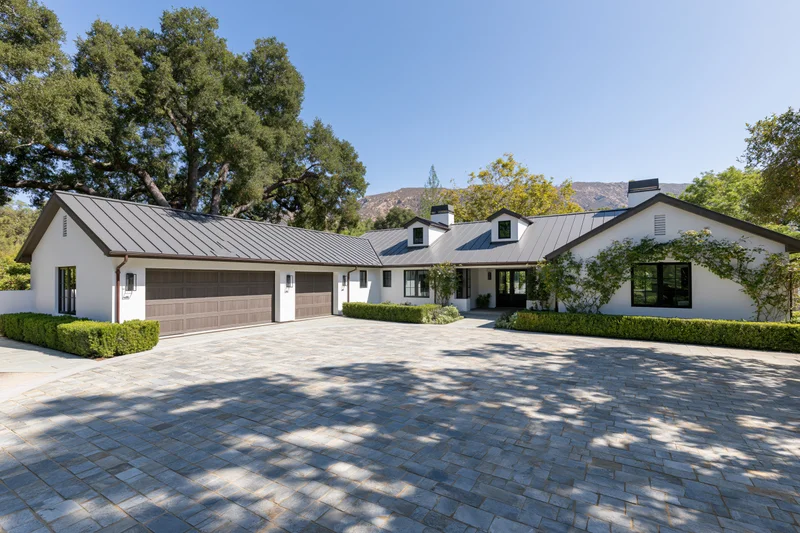 Modern white stucco farmhouse with dark metal roof and stone driveway surrounded by trees and hills