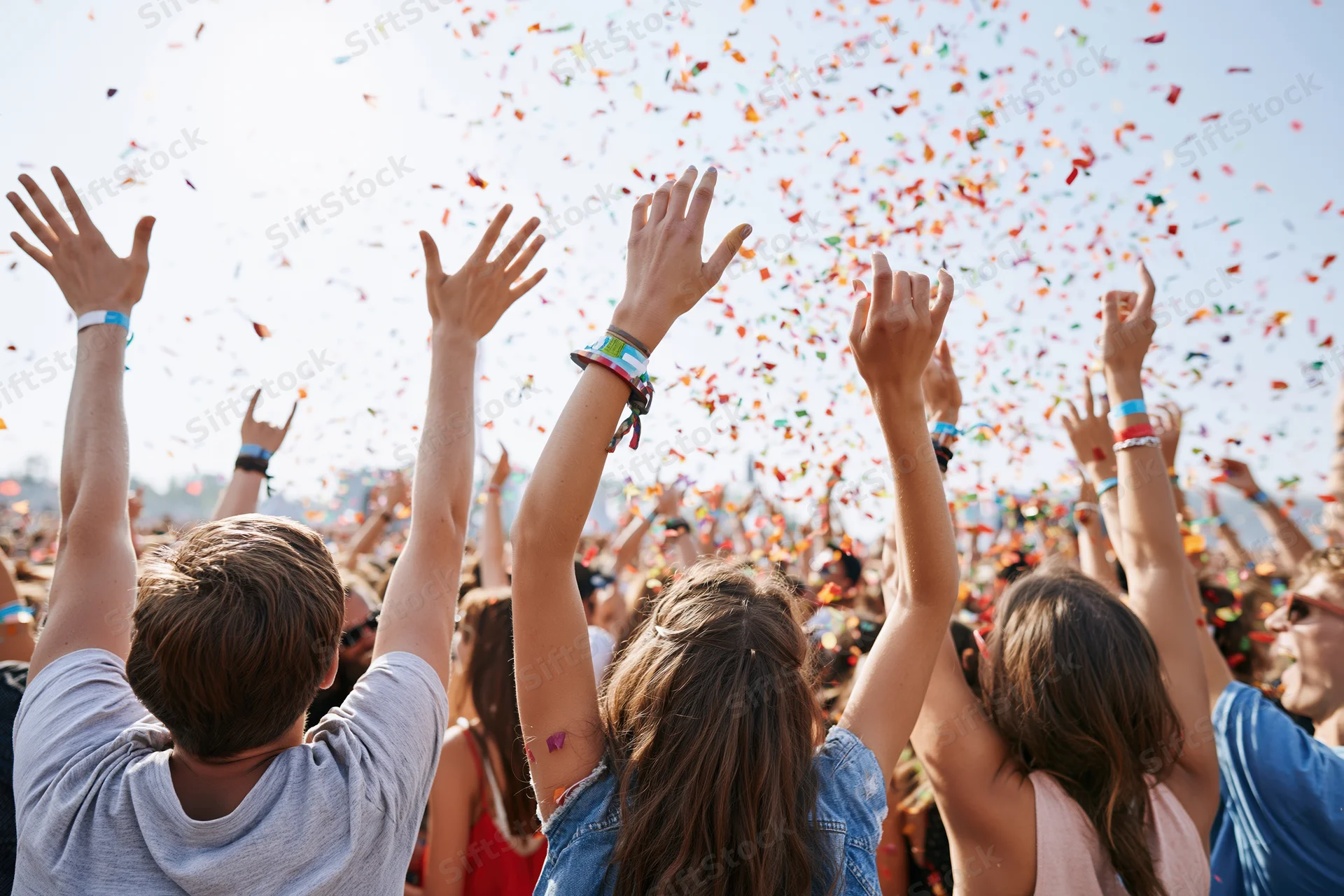 Crowd of young adults with arms raised under falling confetti at outdoor festival