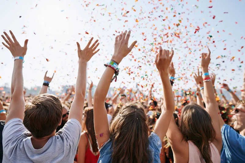 Crowd of young adults with arms raised under falling confetti at outdoor festival