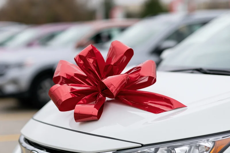 Large shiny red gift bow on the hood of a white car in a dealership lot