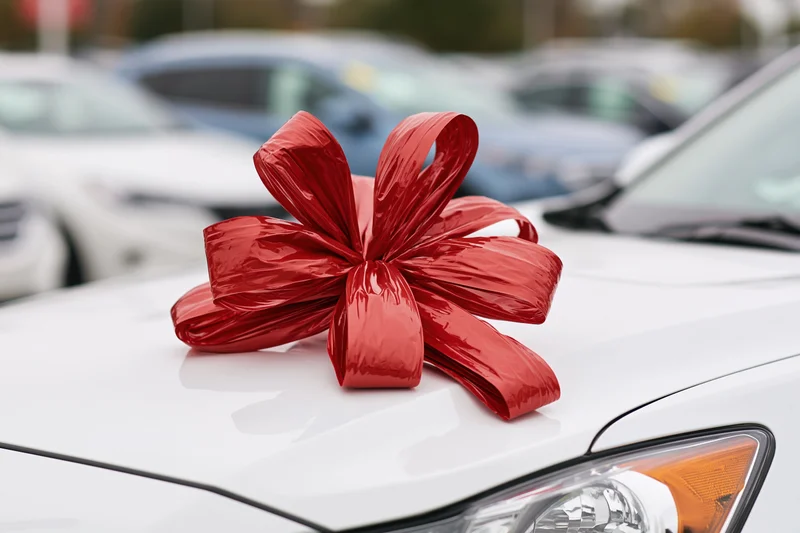 Large shiny red gift bow on the hood of a white car in a parking lot