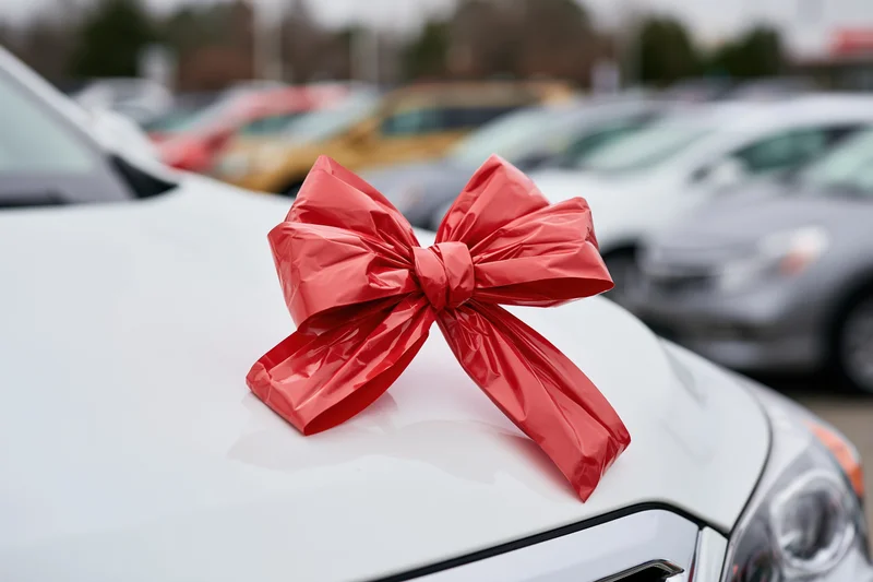 Large shiny red gift bow on the hood of a white car in a dealership lot