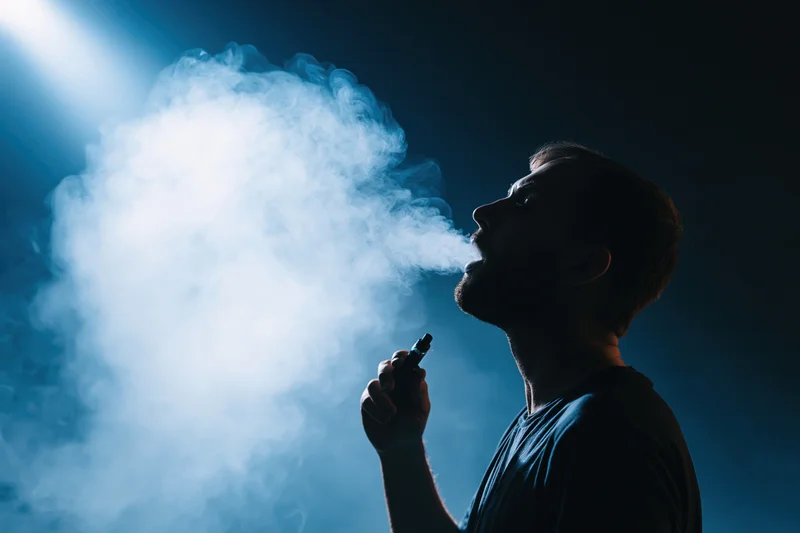 Man exhaling vapor from e-cigarette in dark blue-lit studio setting