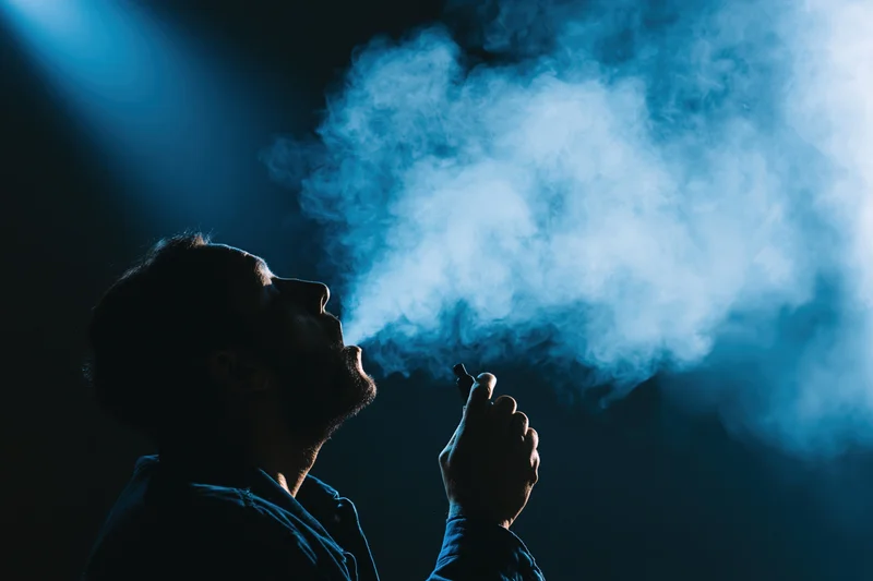 Man exhaling vapor cloud while holding vaping device in dark blue-lit setting