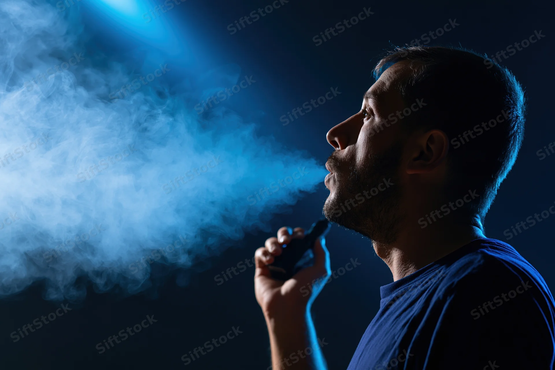 Man exhaling vapor from handheld device in dark blue-lit studio setting