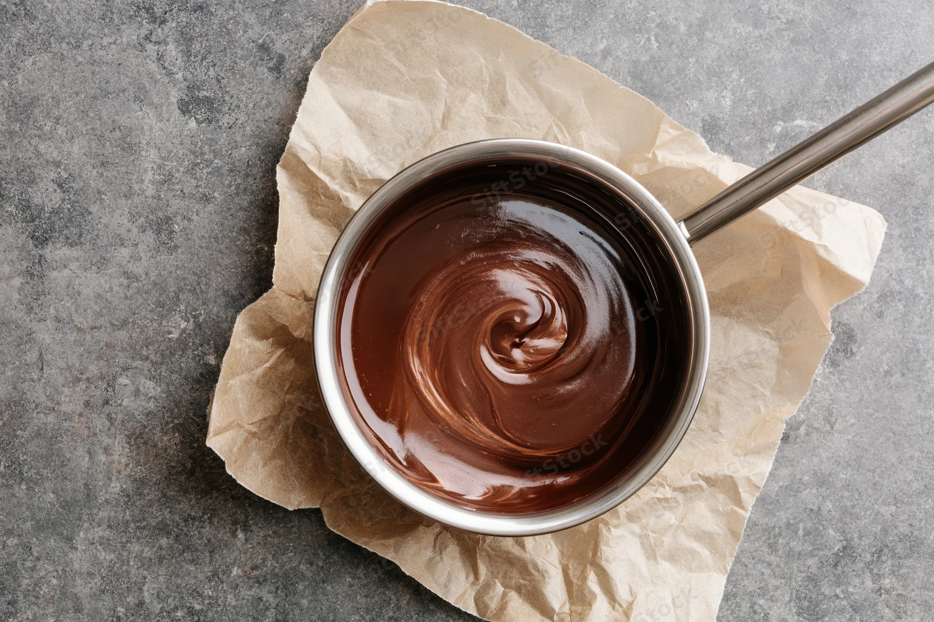 Melted chocolate swirling in a small stainless steel saucepan on crumpled parchment paper over gray stone surface