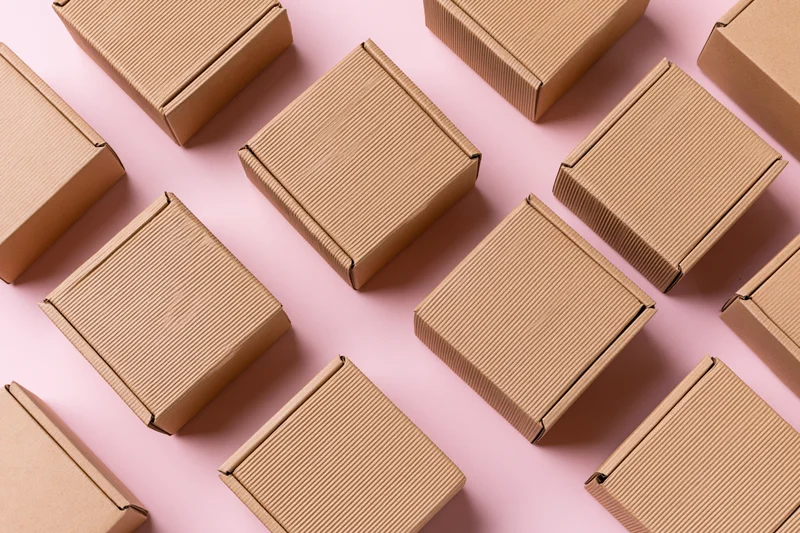 Multiple small closed kraft cardboard mailer boxes arranged in a pattern on a pink surface