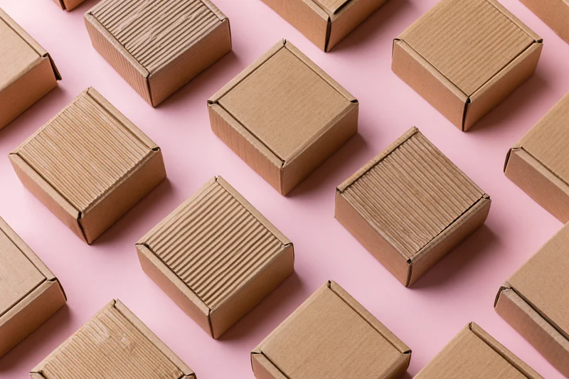 Multiple small closed kraft cardboard mailer boxes arranged in a flat lay on a pink background