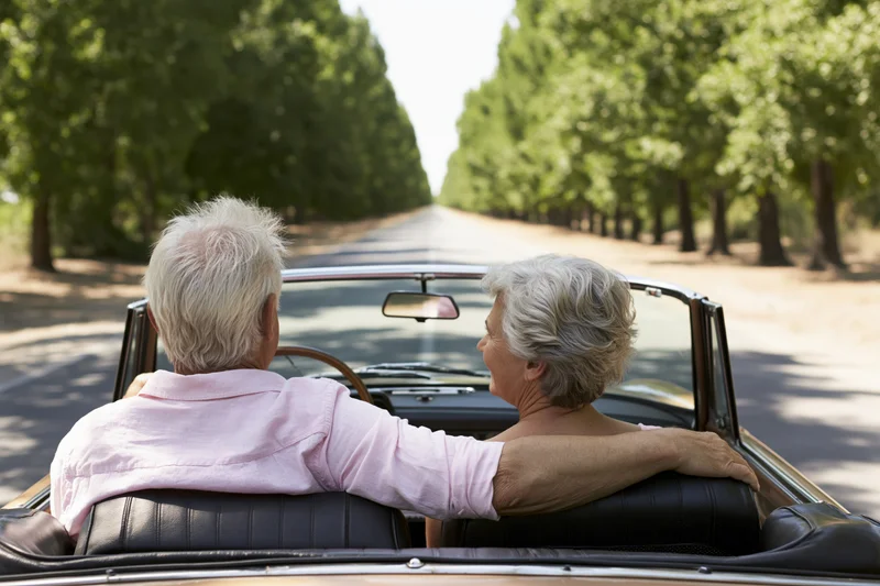 Senior couple driving vintage convertible on tree-lined road, seen from behind