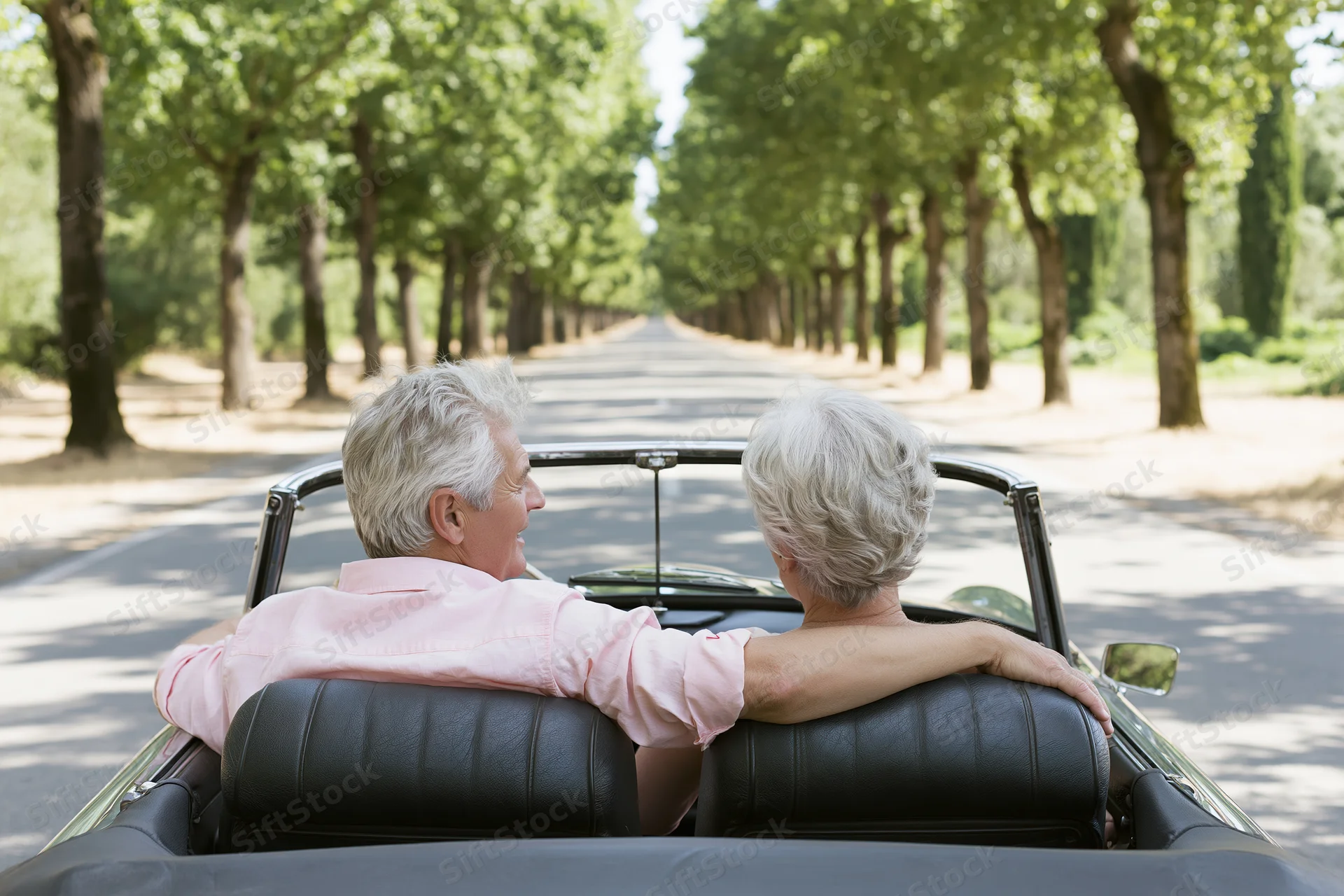 Senior couple driving vintage convertible on tree-lined road, seen from behind, smiling and leaning on each other