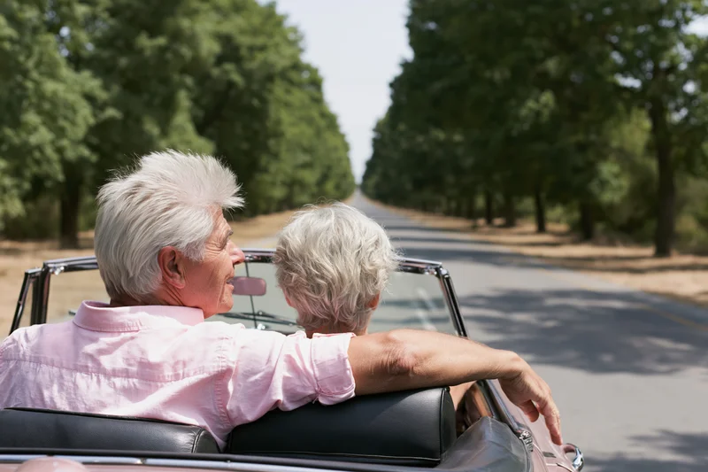 Senior couple driving vintage convertible on tree-lined road in summer