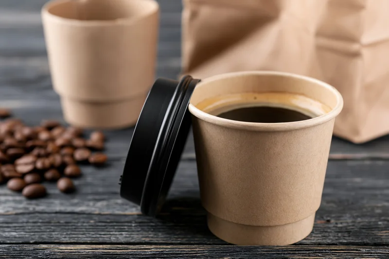 Open kraft paper coffee cup filled with dark espresso on a wooden table with coffee beans and a paper bag nearby