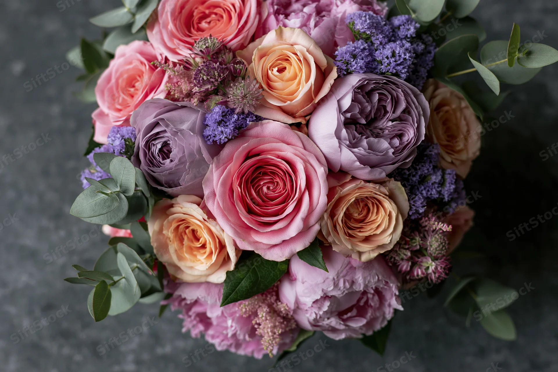 Overhead close-up of a lush mixed flower bouquet with pink roses, purple peonies, lavender, and eucalyptus leaves on a dark textured surface