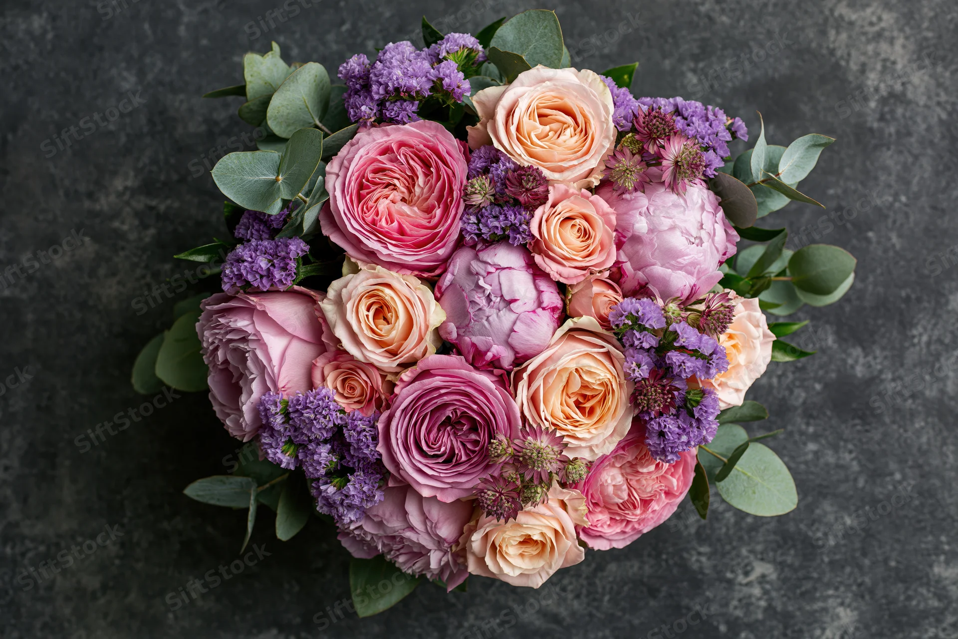Overhead close-up of a lush mixed flower bouquet with pink roses, purple lavender, and green eucalyptus on dark textured surface
