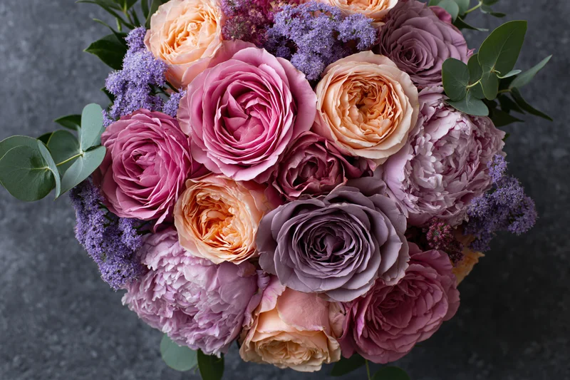 Overhead close-up of a lush mixed flower bouquet with pink roses, purple lilac, peach peonies and green eucalyptus leaves on a dark surface