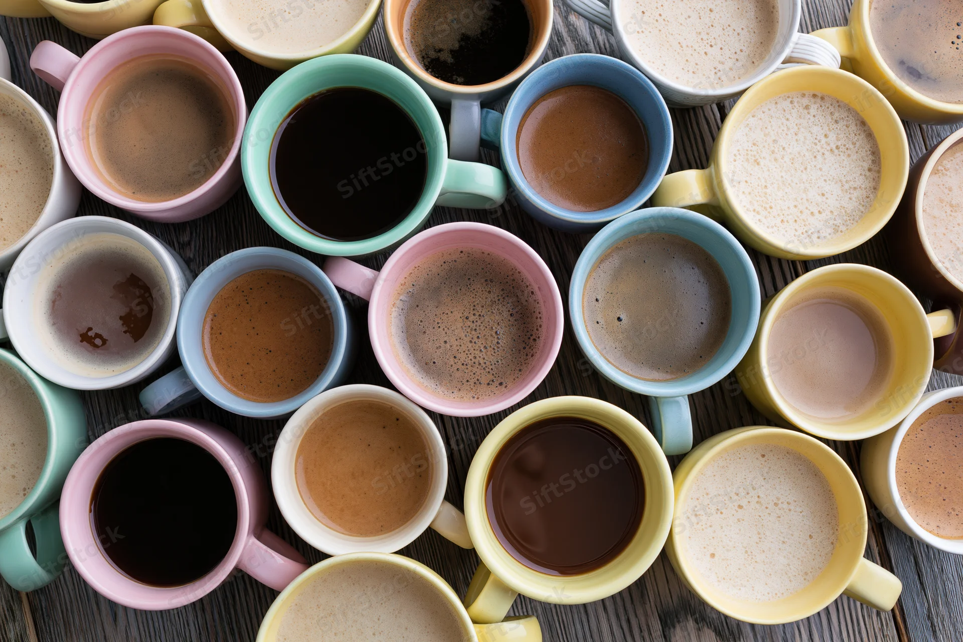 Overhead flat-lay of many mismatched ceramic coffee mugs filled with various coffee drinks on a wooden table