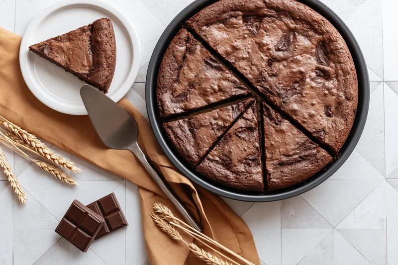 Chocolate brownie cake sliced in pan with one piece on plate and wheat stalks nearby