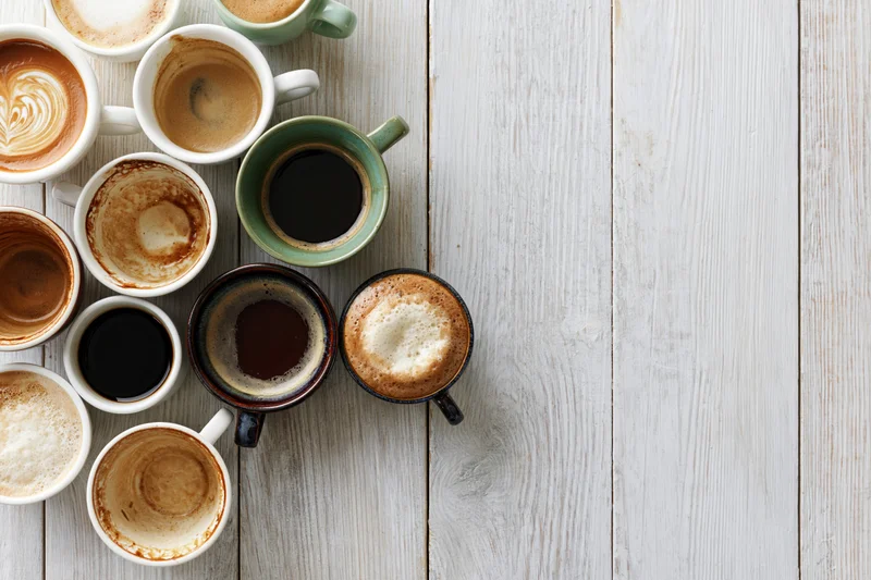 Overhead flat lay of assorted coffee cups with varying contents and residues arranged on a white wooden table