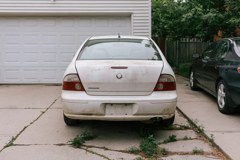 Rear view of a dirty white sedan parked on a cracked concrete driveway in front of a garage